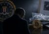 Ehemaliger FBI-Agent sitzt mit gesenktem Kopf vor Bitcoin, Hardware-Wallet und Ermittlungsunterlagen als Symbolbild für den mutmaßlichen Diebstahl von Kryptowährungen.