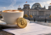 Cappuccino und goldene Bitcoin-Münze auf symbolischen Steuerunterlagen vor dem Berliner Reichstagsgebäude.