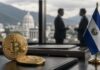 Bitcoin-Münzen und El-Salvador-Flagge auf einem Schreibtisch in einem Büro, mit verschwommenen Geschäftsleuten im Hintergrund