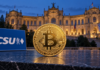 Goldene Bitcoin-Münze neben einem CSU-Logo vor dem beleuchteten Maximilianeum in München.