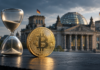 Krypto-Haltefrist: CDU-Mann Güntzler nimmt Klingbeils Steuerpläne auseinander Goldene Bitcoin-Münze neben einer Sanduhr vor dem Reichstagsgebäude in Berlin.