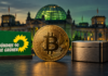 Goldene Bitcoin-Münze neben einer metallenen Spardose vor dem unscharfen Reichstagsgebäude mit grün beleuchteter Kuppel.