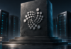 Silbernes IOTA-Symbol auf einem erhöhten dunklen Sockel vor Serveranlagen.