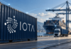 IOTA im Welthandel: Ericsson, Scania, H&M und weitere Giganten an Bord Container mit großem IOTA-Logo vor einem Frachtschiff und einem Lkw, verbunden durch symbolische digitale Datenlinien.