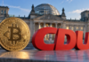 Goldene Bitcoin-Münze neben einem roten CDU-Logo vor dem Reichstagsgebäude in Berlin.