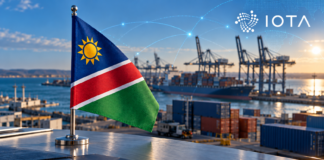 Wird IOTAs ADAPT auf Namibia ausgeweitet? Was bislang bekannt ist Namibische Flagge vor einem Containerhafen mit digitalen Verbindungslinien und IOTA-Logo.